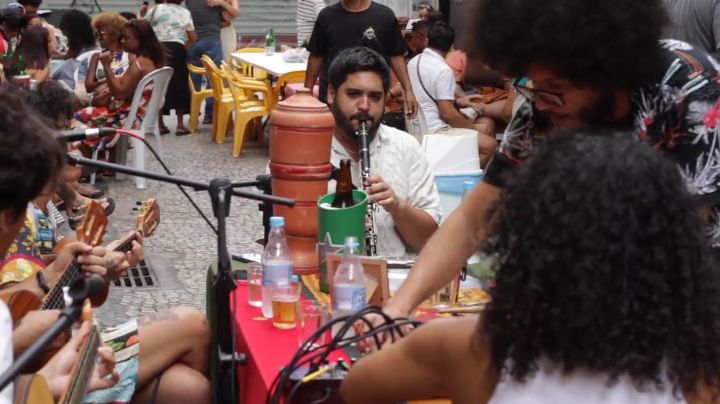 A imagem mostra uma roda de samba ao ar livre, com músicos tocando instrumentos de corda e sopro cercados por mesas e pessoas sentadas na calçada. O ambiente é movimentado e informal, típico de um encontro de boteco no Centro de Vitória.