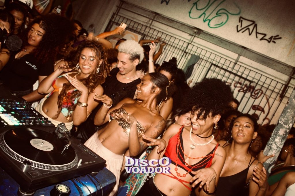 Grupo de pessoas dançando animadamente ao redor de uma cabine de DJ em uma festa noturna. Em primeiro plano, um toca-discos com vinil e uma mesa de mixagem iluminada aparecem no canto esquerdo. Ao centro, várias pessoas sorriem e dançam com os braços levantados, usando roupas coloridas, acessórios brilhantes e penteados volumosos. Ao fundo há grades metálicas e grafites na parede, iluminados por luz quente de balada. No centro inferior da imagem aparece o texto ‘Disco Voador