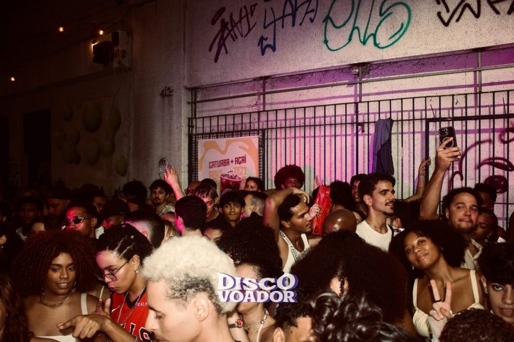 Multidão reunida em uma festa de rua noturna, em frente a um prédio com grades e paredes cobertas de grafites. Diversas pessoas aparecem dançando, conversando e usando celulares, iluminadas por luzes rosadas e quentes. A cena transmite clima animado e cheio de movimento, com pessoas de diferentes estilos e aparências ocupando quase toda a imagem. No centro inferior aparece o texto ‘Disco Voador