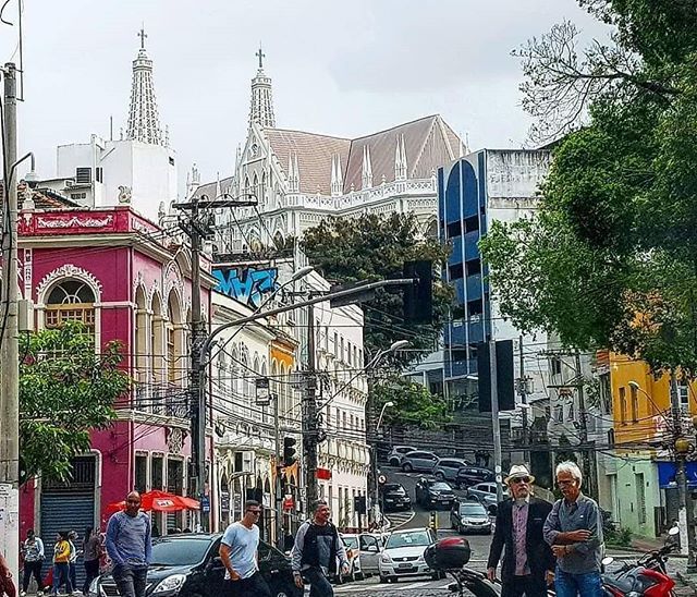 Vista de uma rua do Centro de Vitória durante o dia. Em primeiro plano, pessoas caminham e atravessam a via enquanto carros e motocicletas circulam pelo trânsito. Nas laterais, edifícios históricos de diferentes cores, como rosa, amarelo e verde, contrastam com construções mais modernas. Ao fundo, em posição elevada, destaca-se a Catedral Metropolitana de Vitória, com suas torres brancas e telhado inclinado, cercada por árvores e edificações. A imagem evidencia a mistura entre patrimônio histórico, movimento urbano e arquitetura característica da região central da cidade.