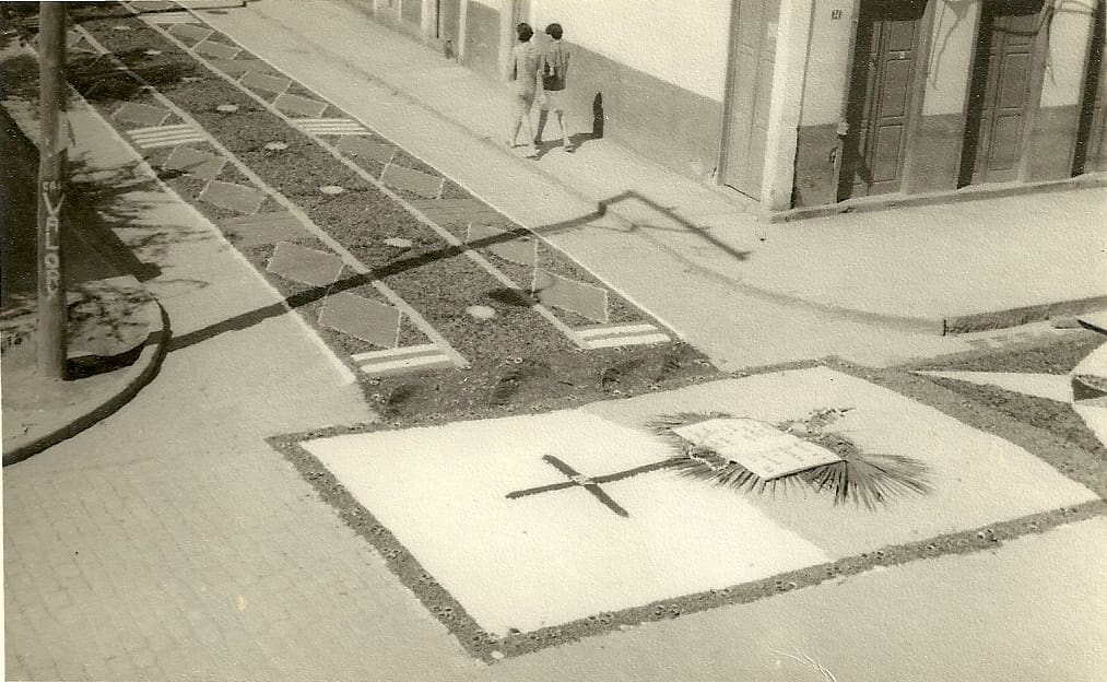 Foto antiga, com coloração sépia, de um dos primeiros tapetes de Corpus Christi confeccionados em Castelo, que continha folhas, cascas de café, flores e areia. A imagem exibida no tapete é de uma cruz, com uma bíblia e um terço logo abaixo, estes em cima de folhas de palmeira, formando uma das partes do tapete, que continua com um padrão que possui losangos ao lado e uma faixa contínua de cascas de café ao centro, formando uma das passadeiras do tapete.