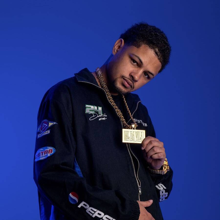 Homem posa para um ensaio fotográfico diante de um fundo azul iluminado. Ele usa jaqueta preta com aplicações e logotipos nas mangas, correntes douradas no pescoço, relógio dourado e segura um grande pingente dourado com uma das mãos. Está levemente inclinado para a frente, olhando diretamente para a câmera com expressão séria. A iluminação destaca o contraste entre as roupas escuras e o fundo azul vibrante.